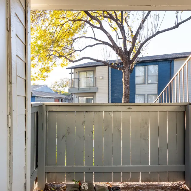 A view from a small balcony with wooden railing, looking out towards a tree and adjacent apartment buildings in the background.