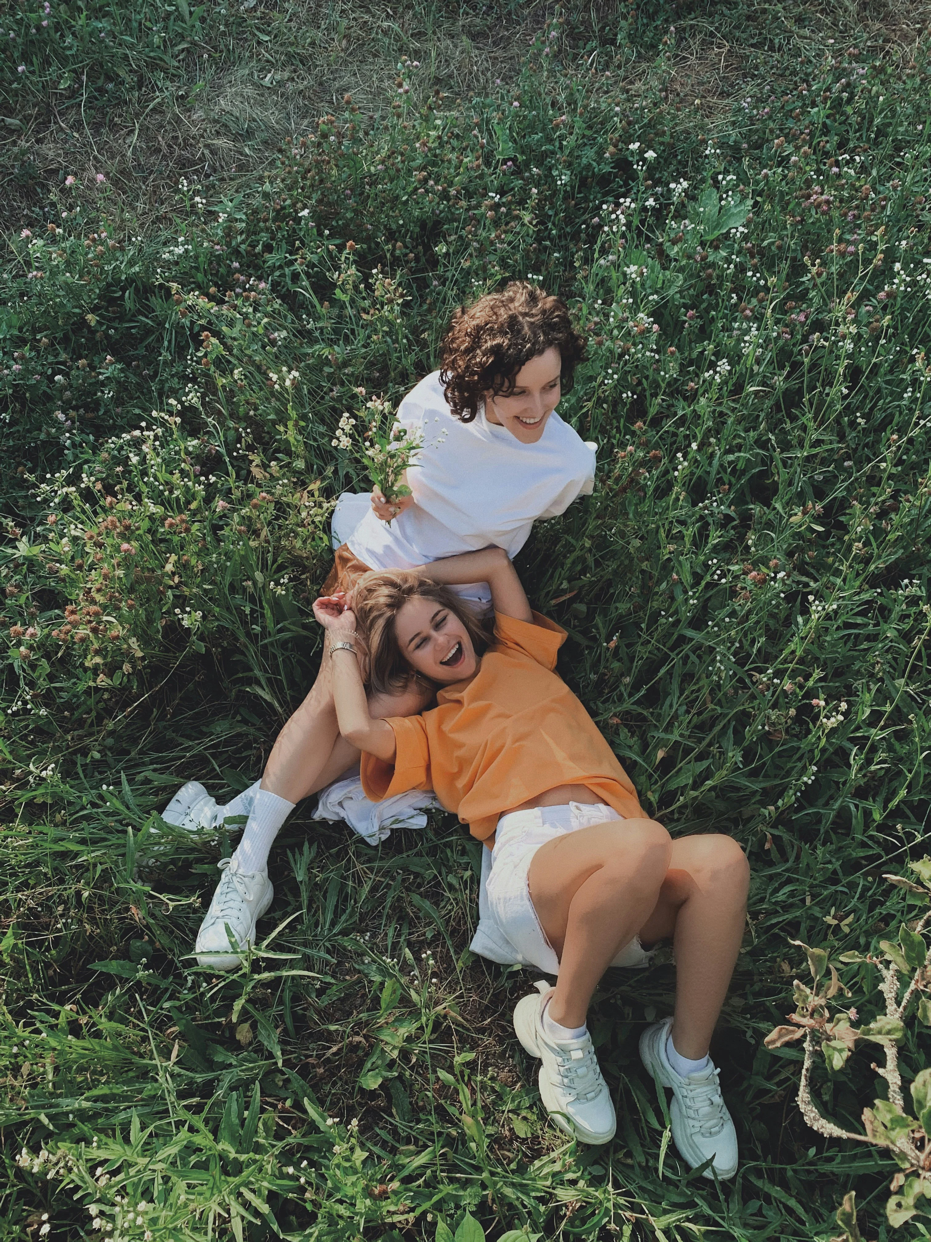 Two young women lounging in a grassy field, one with curly hair in a white shirt and the other with straight hair in an orange shirt. They appear happy and relaxed, surrounded by greenery and flowers.