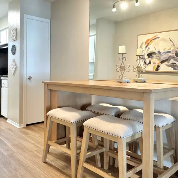 A modern dining area featuring a wooden table with cream-colored padded stools, a rhythmic abstract painting on the wall, and a well-lit kitchen space in the background.