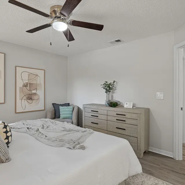 A cozy modern bedroom featuring a white bed with decorative pillows, a wooden dresser, and abstract art on the walls. A ceiling fan is installed, and there is a glimpse of a closet in the background.