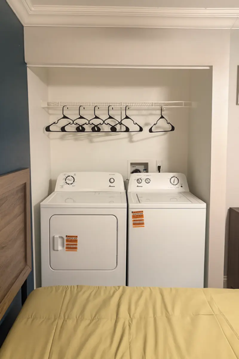 Laundry Area in Room A compact laundry area featuring a washing machine and dryer side by side, with a shelf above holding several black hangers. The laundry appliances have labels attached and are located against a wall in a small room, next to a bed with a yellow bedspread.