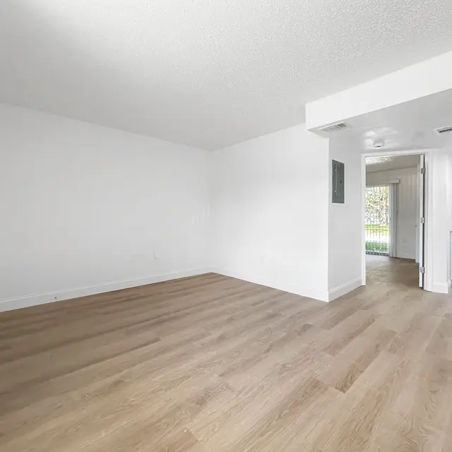 Spacious Empty Room A spacious and empty room with light-colored walls and wooden flooring, featuring a doorway leading to a small kitchen area.