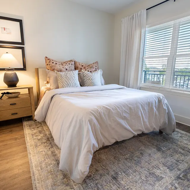 A cozy bedroom featuring a comfortable bed, decorative pillows, and a large window with blinds letting in natural light. There's a wooden nightstand with a lamp and decor items.