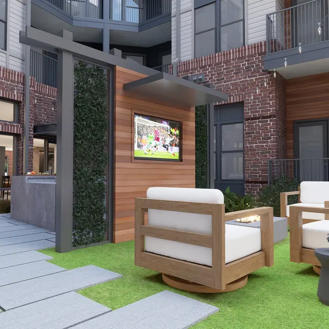 A modern outdoor lounge area with two white armchairs on a green lawn, a small side table, and a wall-mounted TV surrounded by greenery, set against a backdrop of brick and modern architecture.