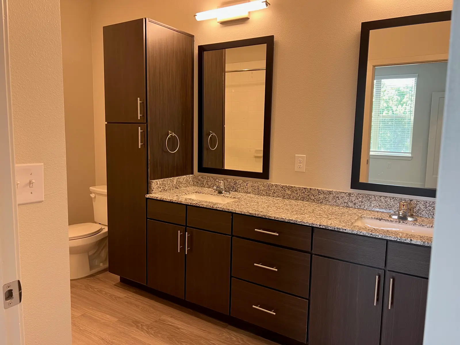 A modern bathroom featuring dark wooden cabinetry, a double sink counter with granite top, two mirrors, and a toilet in the corner. Natural light is coming through a window at the back.