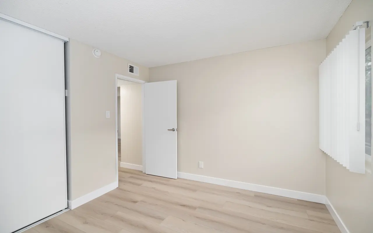 A spacious, empty room with light-colored walls and wood-style flooring. There is a sliding closet door on the left and a door leading to another area on the back wall. Natural light filters in through a window with blinds on the right side.