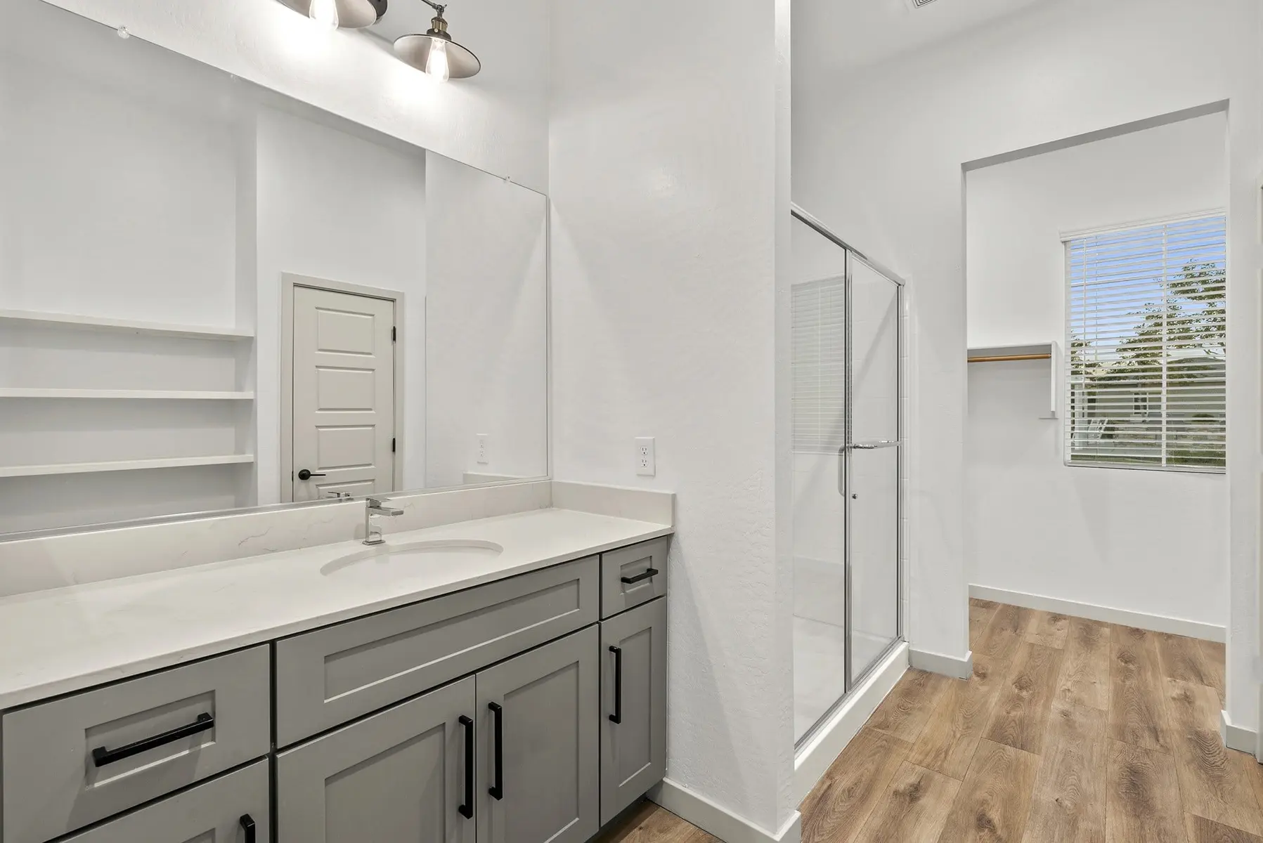 Modern bathroom with a double vanity, shower stall, and large window, featuring light-colored walls and wooden floor.