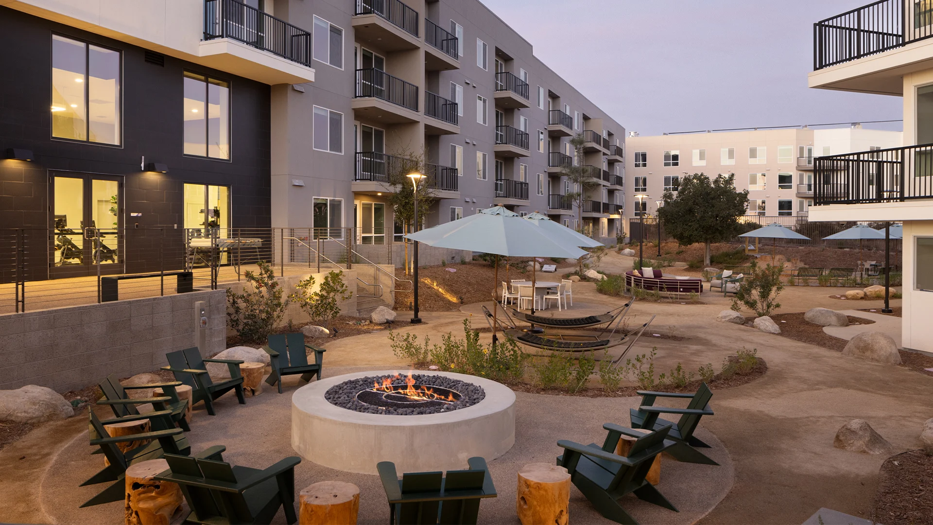 A spacious outdoor living area featuring a fire pit surrounded by wooden chairs, with multiple apartment buildings in the background, and landscaped pathways.