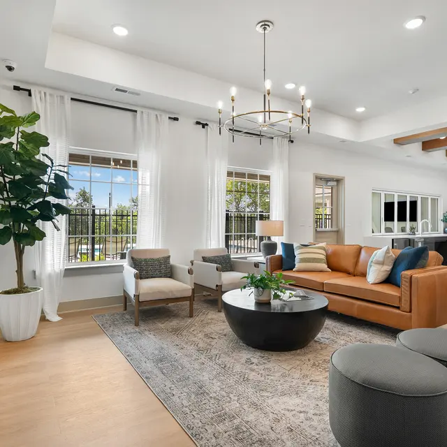 A modern living room featuring a brown sofa, light-colored armchairs, and decorative plants. The room has large windows allowing natural light, with stylish light fixtures and a flat-screen TV on the wall. A circular coffee table and round ottomans complete the setup.