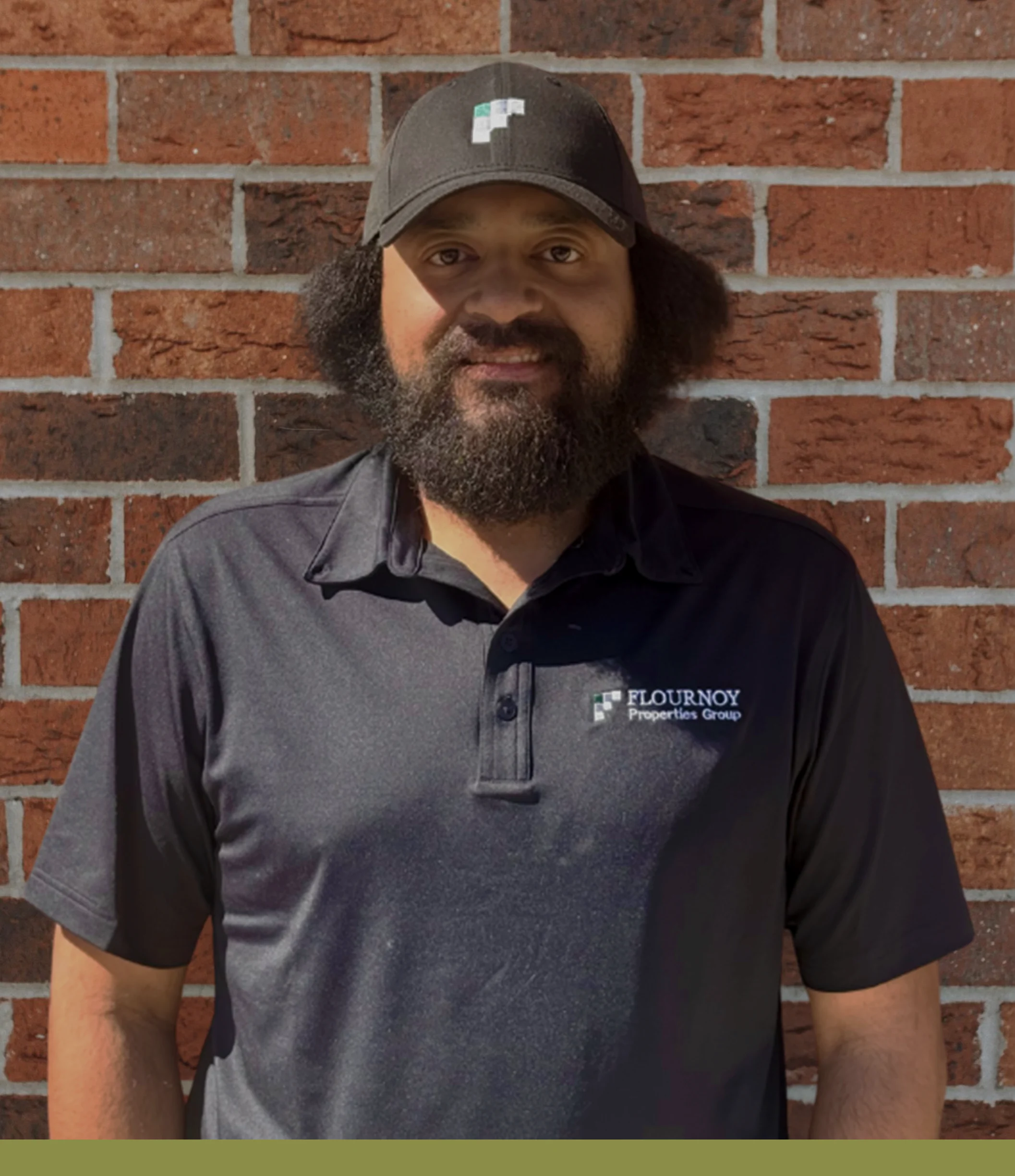 Man in Branded Attire A man standing in front of a brick wall, wearing a black shirt with a logo and a matching hat.