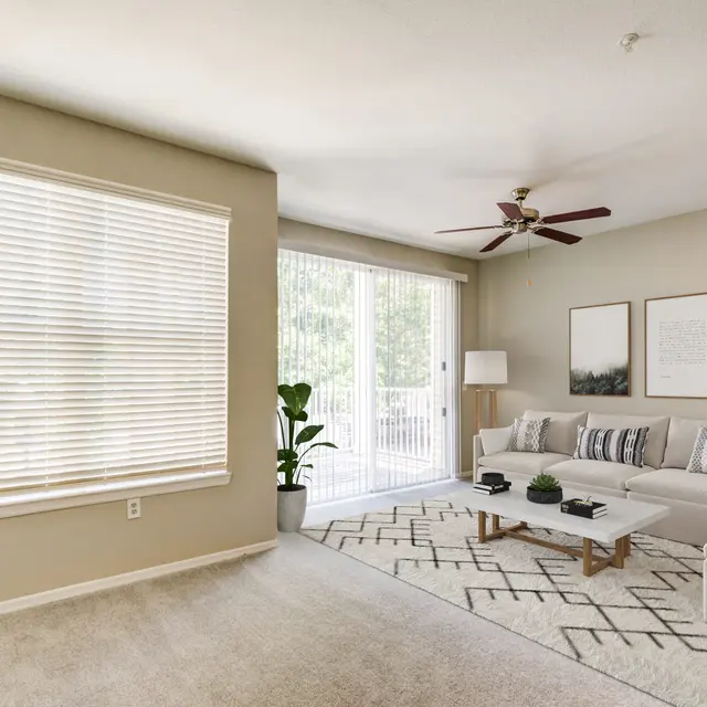 A stylish modern living room featuring a beige color scheme, a large window with blinds, a comfortable couch with decorative cushions, a coffee table with books and plants, and a light rug with a geometric pattern.