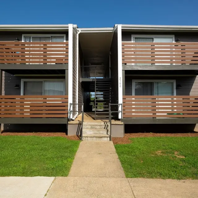 Exterior view of an apartment complex featuring two levels with wooden balcony railings and a central staircase.