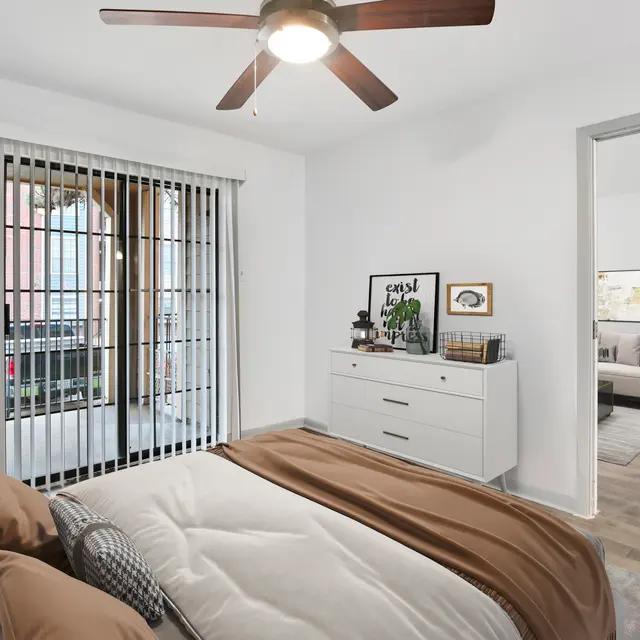 A modern bedroom featuring a bed with brown and white bedding, a fan on the ceiling, a dresser with decor, and a sliding door that opens to an outdoor view with vertical bars.