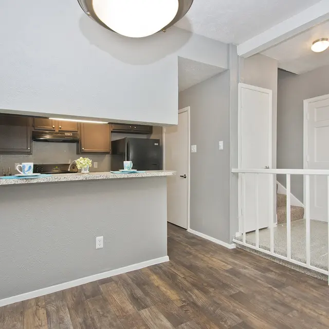 A modern apartment interior featuring an open layout. The area includes a kitchen bar with granite countertop, and adjacent to it is a well-lit entrance with a small staircase leading upwards.