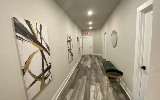 A modern hallway with light gray walls, wooden flooring, and decorative art pieces on the walls. It features a green bench with a gold-colored accent table and a round mirror.