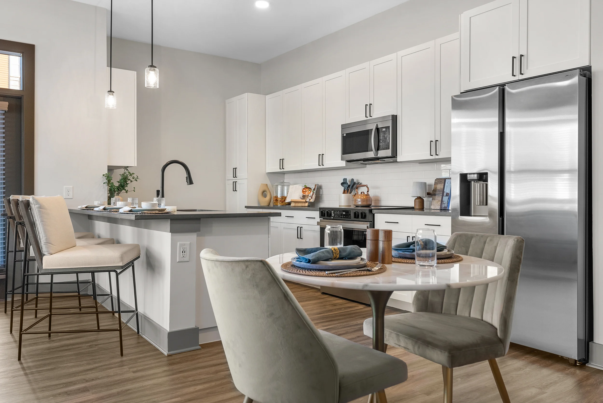 A modern kitchen featuring white cabinets, stainless steel appliances, and a circular dining table with four chairs. There are bar stools at a kitchen island, and the space is bright with natural light.