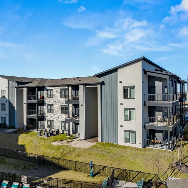 Exterior view of a modern apartment complex featuring two buildings with gray and stone facades, surrounded by green space and fencing.