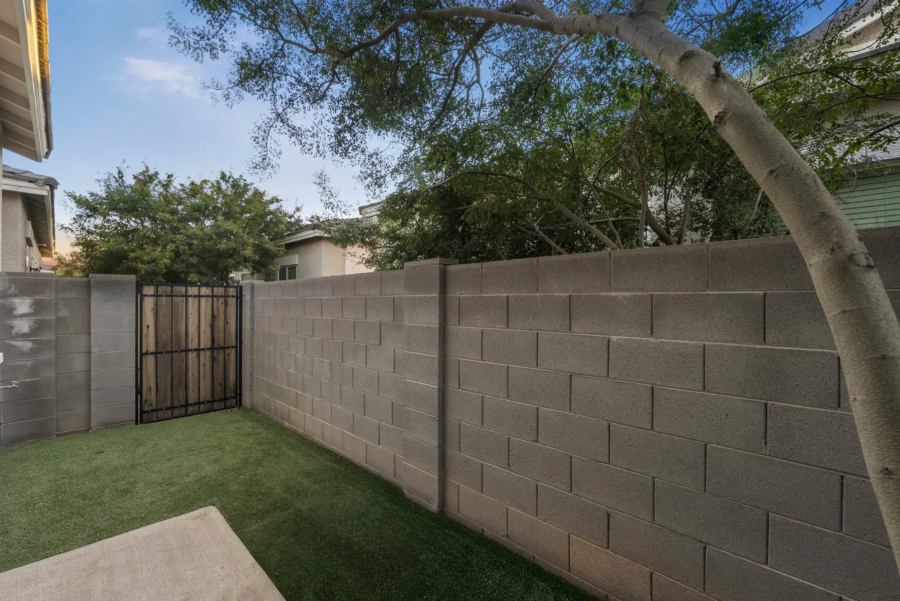 A fenced yard with a grassy area, surrounded by walls and trees, featuring a gate leading to another section.