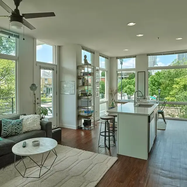 A modern open-concept living area featuring a gray sectional sofa, a round coffee table, and a kitchen with bright white cabinets. Large windows let in natural light and show green trees outside.