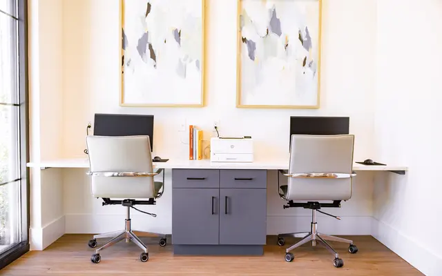 A stylish modern office with two computer desks, each having a monitor and ergonomic chairs. The setup is next to large windows and features abstract artwork on the walls.