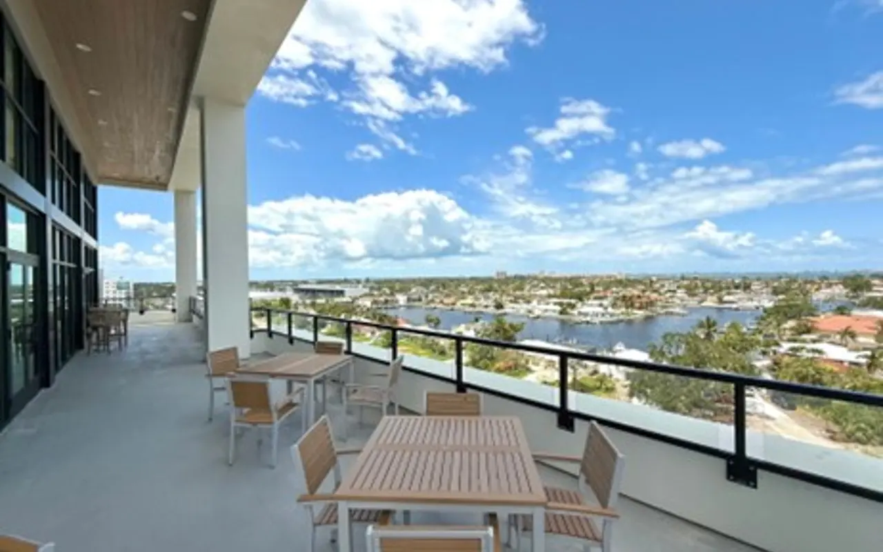 A spacious balcony with wooden tables and chairs overlooking a scenic waterfront view on a sunny day.
