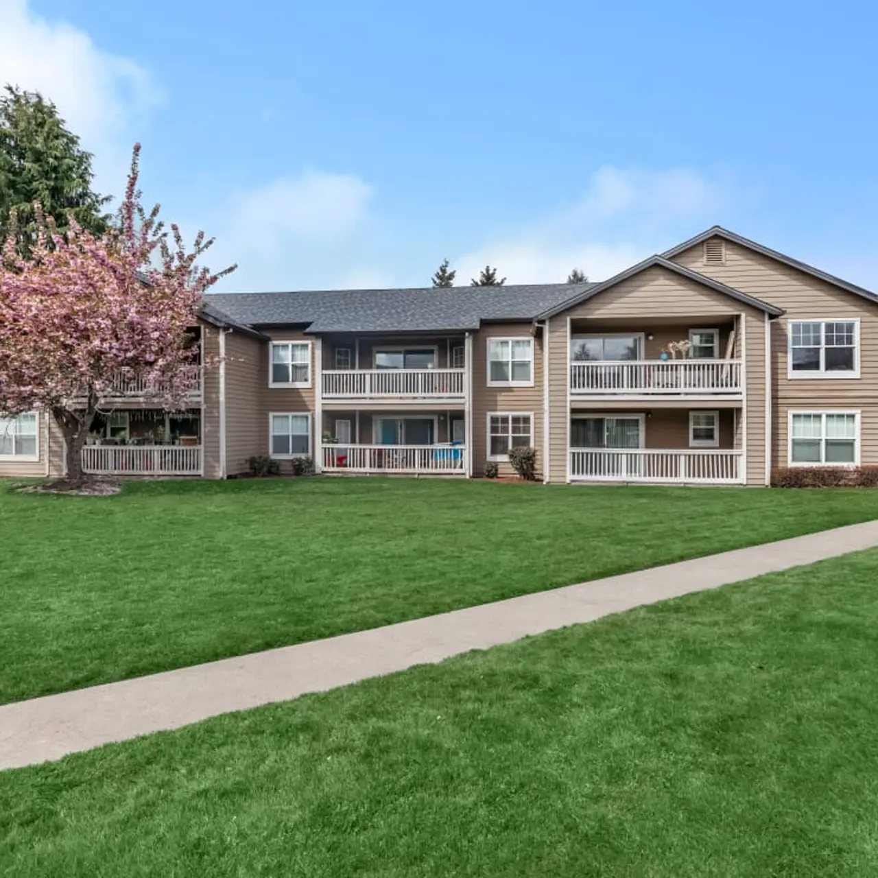 A landscaped apartment complex featuring multiple buildings with balconies, green lawns, and a flowering tree in front.