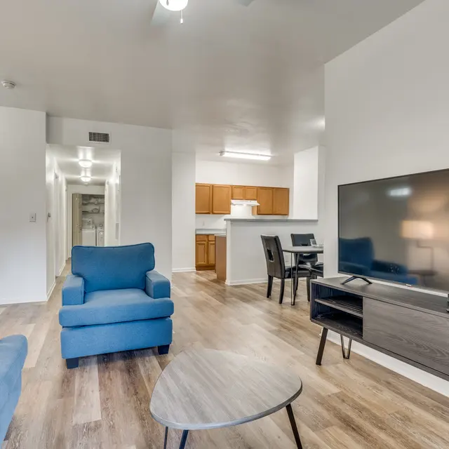 A modern living room featuring blue chairs, a round coffee table, a large TV mounted on the wall, and a kitchen area in the background.