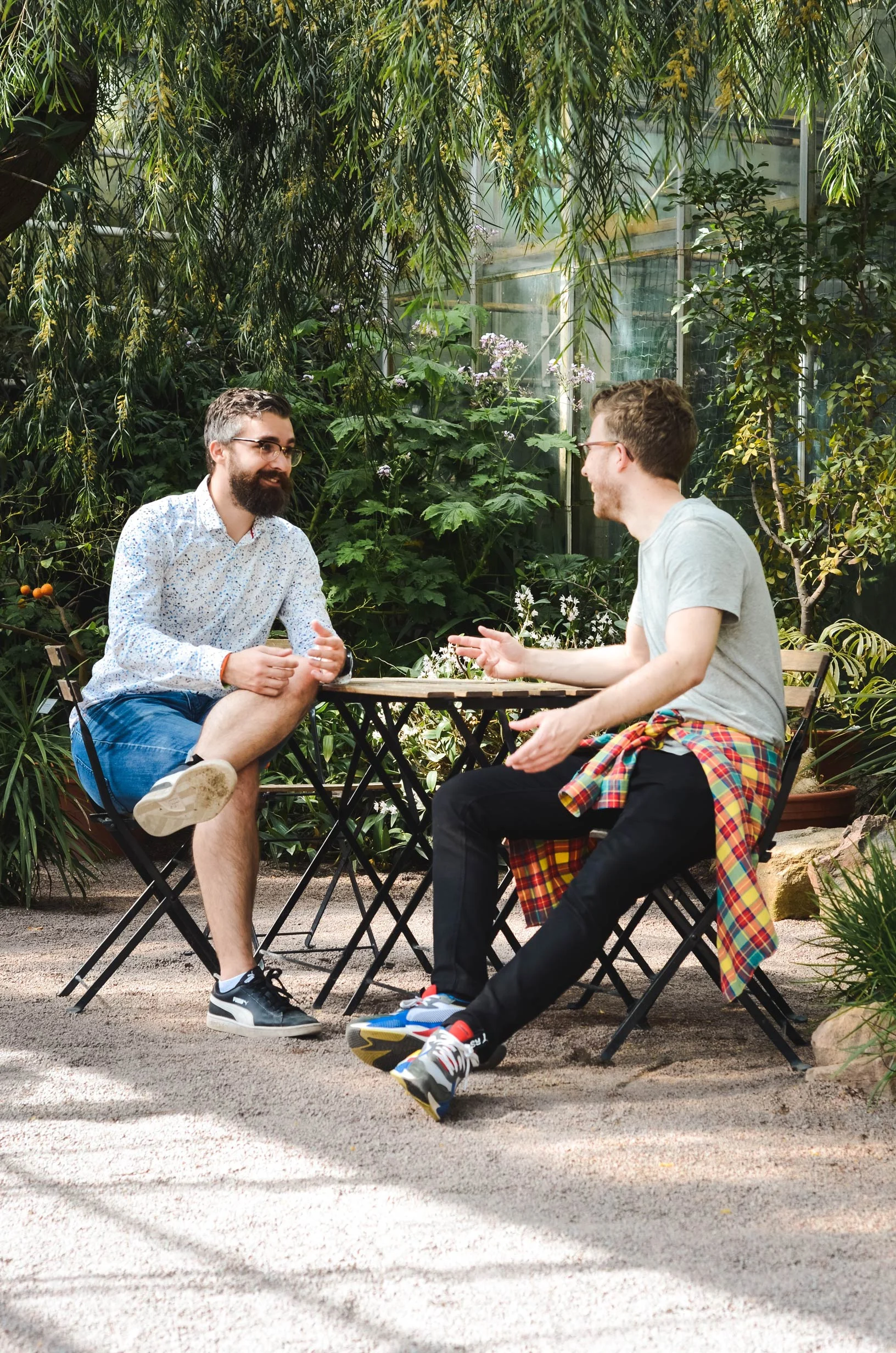 Two men sitting at a small table in a garden cafe area surrounded by greenery. One man has a beard and is wearing a patterned shirt and shorts, while the other is in a light shirt with a plaid shirt tied around his waist. They are engaged in conversation with drinks on the table.