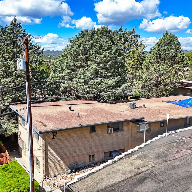 Aerial view of residential buildings with flat roofs and trees in the background under a blue sky with fluffy clouds.