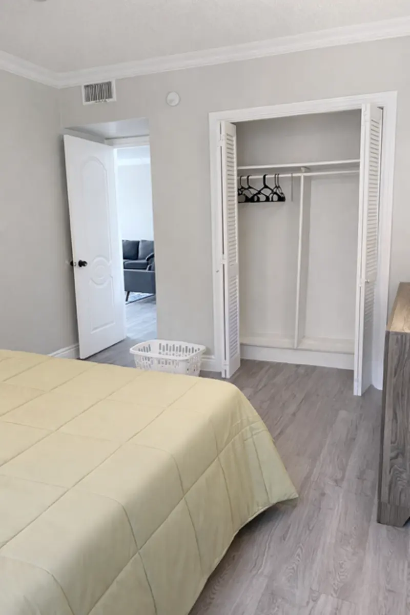 A bedroom featuring a light-colored quilted bedspread, a closet with hangers, and a separate room visible in the background.