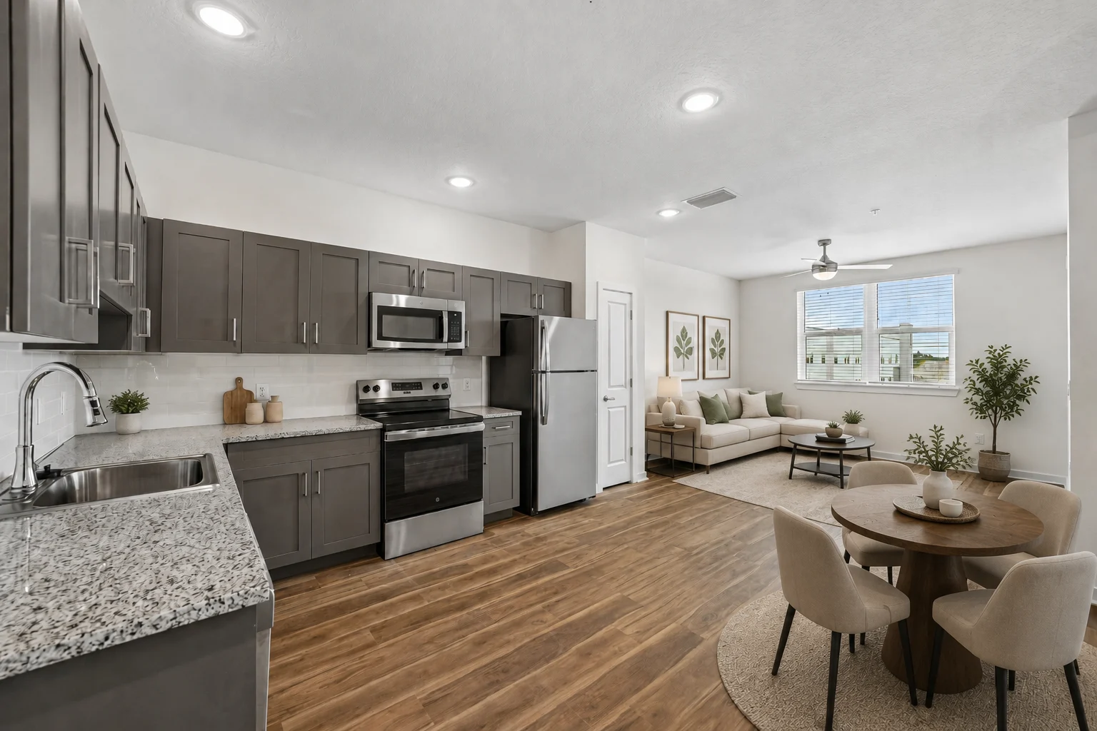 A modern kitchen with gray cabinets, stainless steel appliances, and granite countertops adjacent to a cozy living area with a light-colored sofa and a dining table.