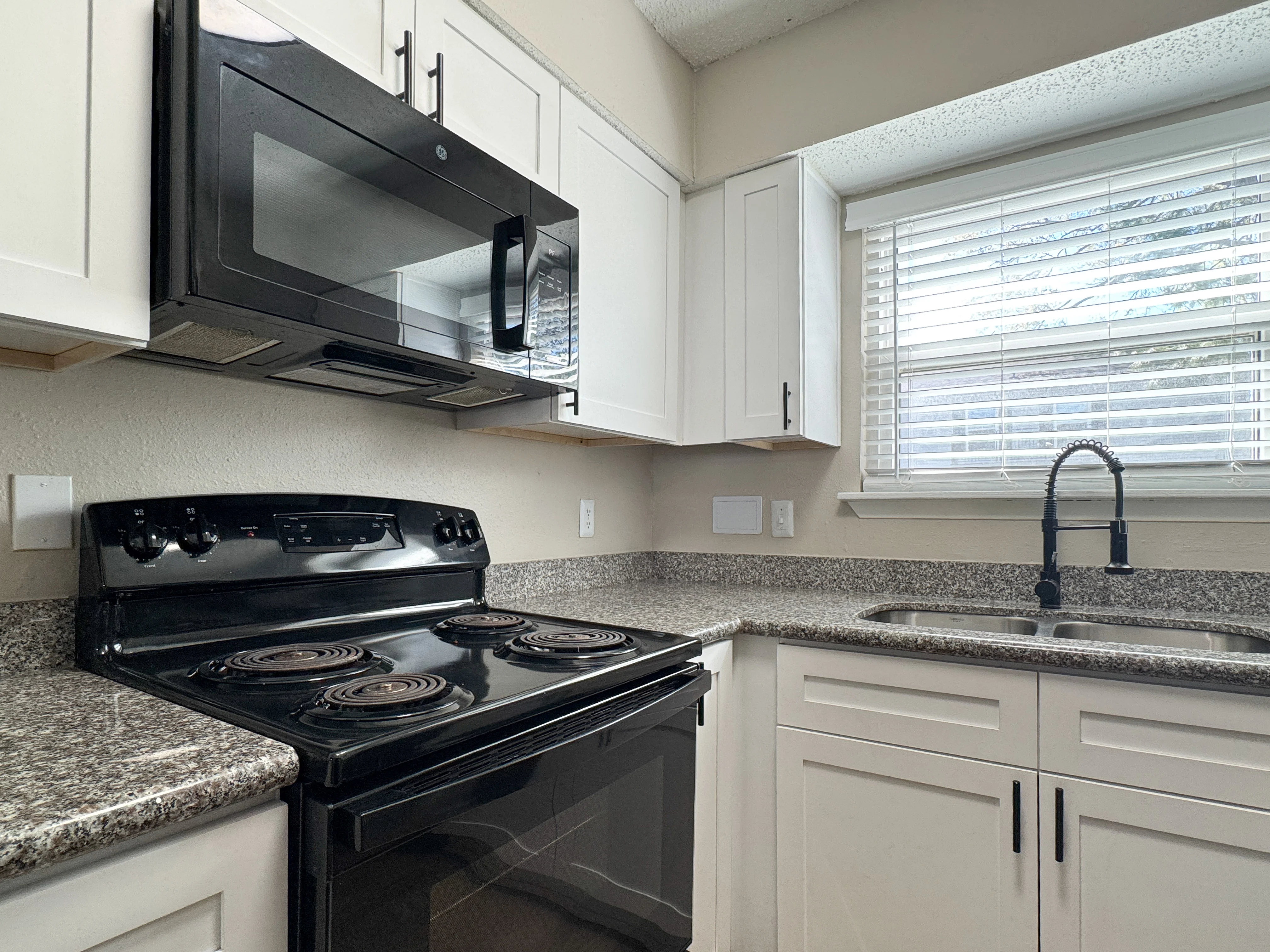 A modern kitchen featuring a black stove and microwave, white cabinets, a granite countertop, and a window with blinds.