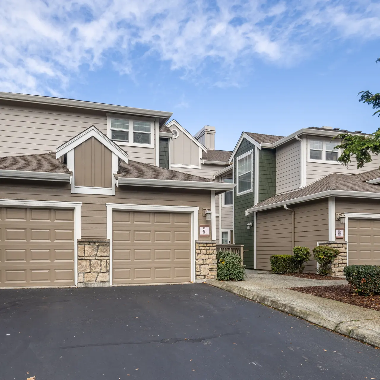 A landscaped residential complex featuring two attached garages and two buildings with a mix of siding colors, set against a partly cloudy sky.