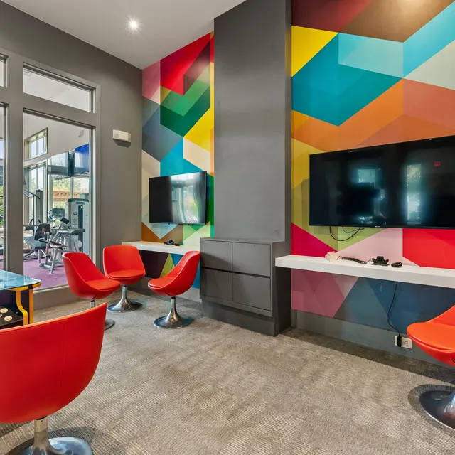 A vibrant game room featuring a geometric patterned wall, two television screens, red chairs, and a game table with controllers.