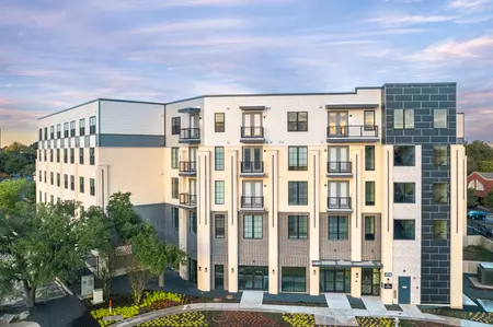 A modern multi-story apartment building with a blend of brick and wood exteriors, featuring large windows and balconies, surrounded by landscaped greenery.