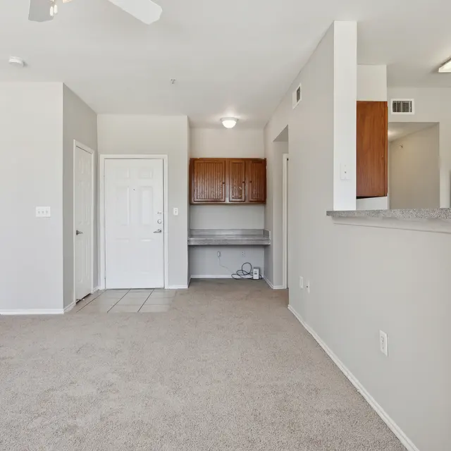 A bright, open living space featuring beige carpet, light walls, and a kitchenette with wooden cabinets and a countertop.