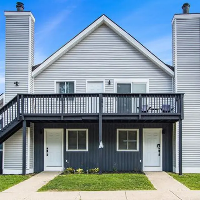 Exterior view of a two-story apartment building with a balcony. Features two front doors and tall chimneys on each side.