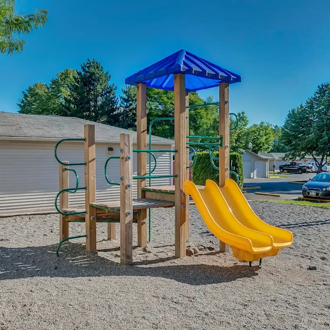 A playground with two yellow slides, a climbing structure, and a sand surface, surrounded by trees and parked cars in the background.