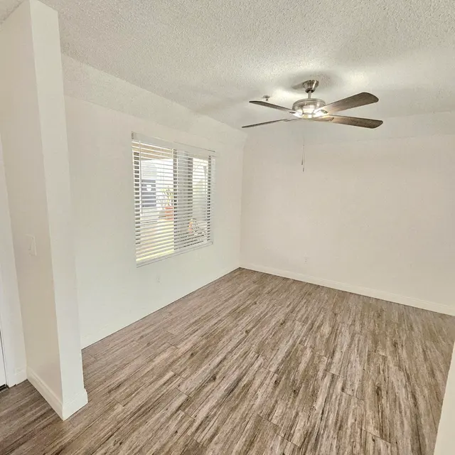 A spacious empty room featuring a ceiling fan, light-colored walls, and wooden flooring. It has two windows with blinds allowing natural light to enter the space.