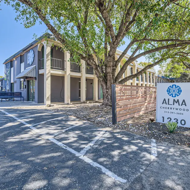 Alma Cherrywood Location Exterior view of Alma Cherrywood building with parking area and a large tree in front, featuring a sign displaying the name and address.
