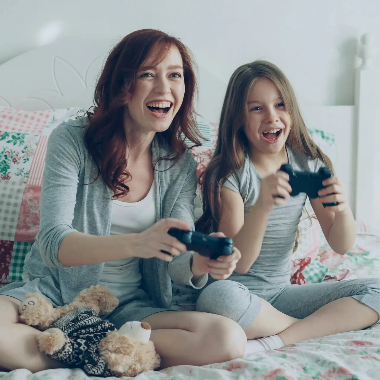 Quality Time: Gaming with Mom A woman and a young girl sitting on a bed playing video games, both smiling and holding game controllers.