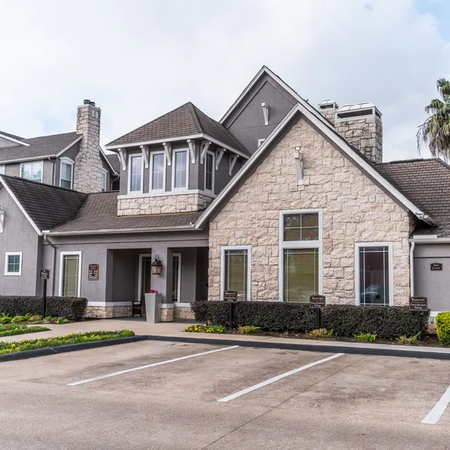 A modern apartment complex with a stone and gray exterior. The building features a peaked roof and several large windows. Surrounding landscaping includes manicured shrubs and palm trees. The parking lot in front has marked spaces and a concrete surface.