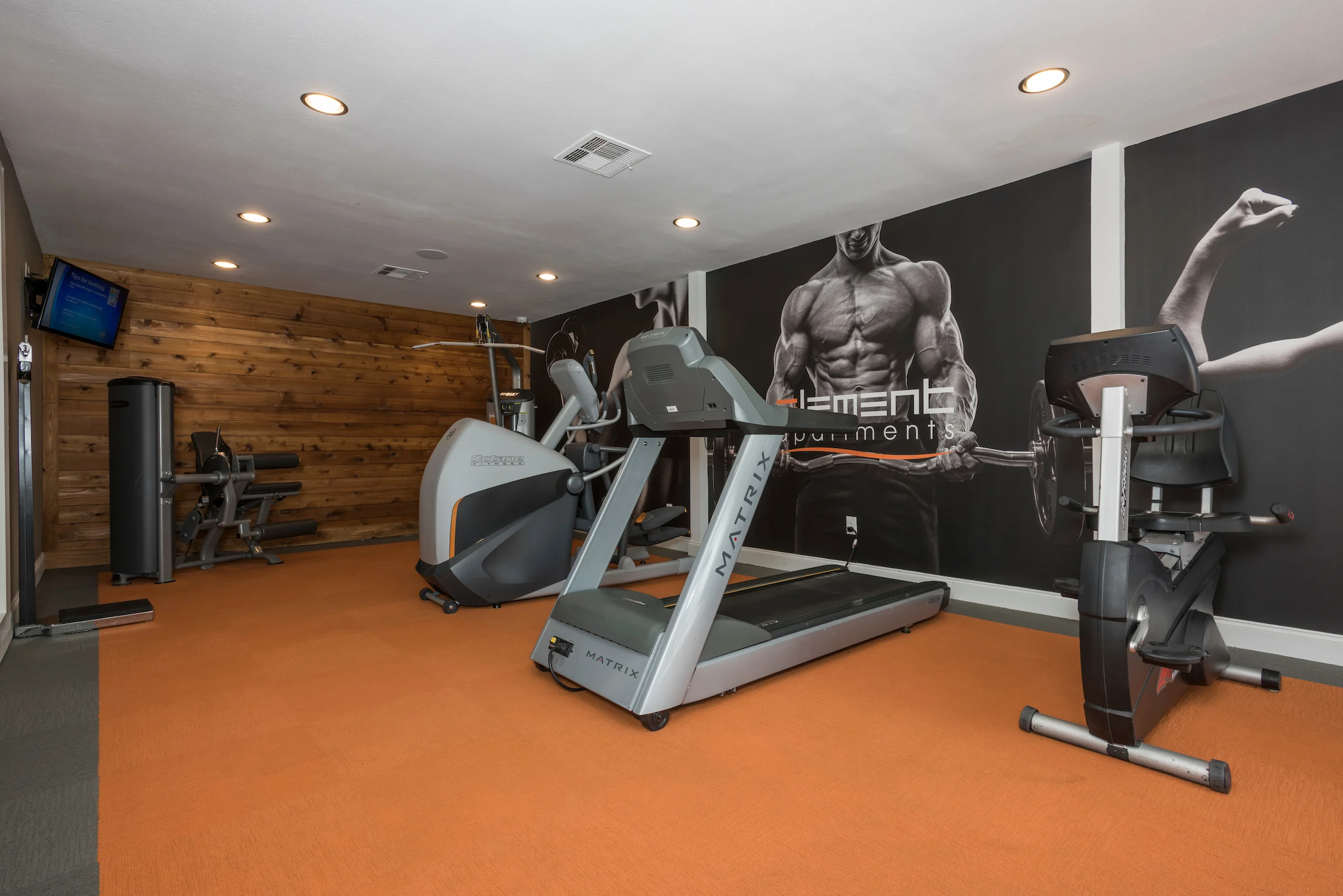 Interior view of a gym with exercise equipment including a treadmill, elliptical, and weight machine, featuring a wooden accent wall and motivational wall art.