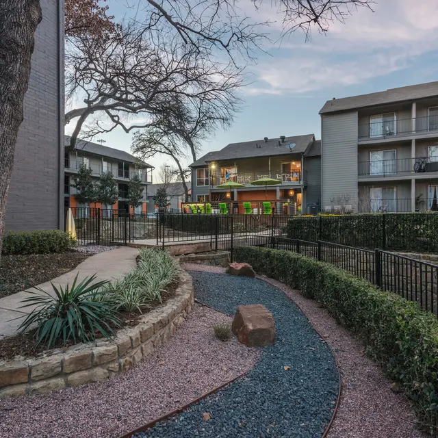 A landscaped apartment courtyard featuring a winding pathway of stones, green shrubs, and several multi-story apartment buildings in the background, with trees and a clear sky visible.