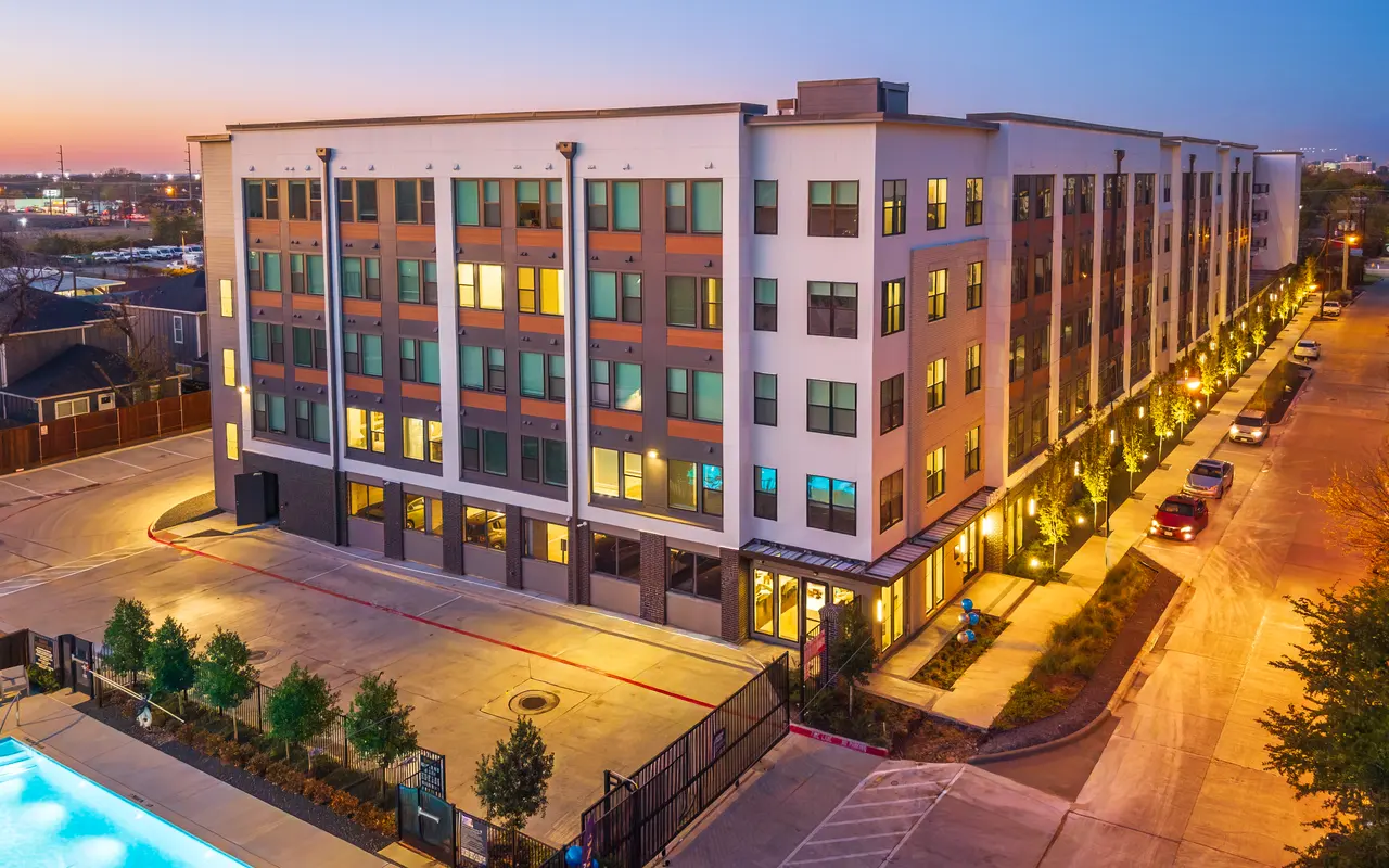 A modern multi-story apartment building illuminated at dusk, with a swimming pool in the foreground and landscaped pathways lined with small trees.