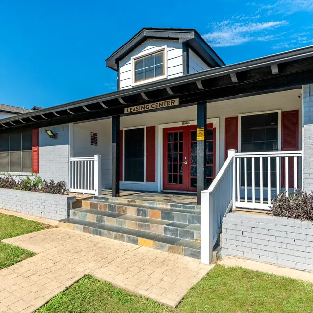 A modern leasing center building with gray brick walls, red shutters, and a sloped roof, surrounded by well-maintained grass and pathways.