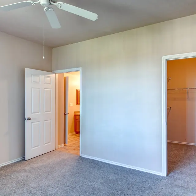 Spacious Empty Room with Doors A spacious room with light beige walls and gray carpet. A ceiling fan is visible, and there are two open doors leading to another room and a closet. The doorway on the left leads to a bathroom.