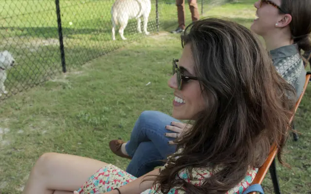 Two women sitting on a bench, laughing and enjoying the view of a dog playing in a fenced area while a man interacts with another dog nearby.