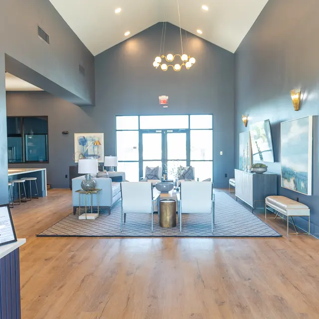 A spacious and modern lobby area featuring high ceilings, contemporary furniture, and large windows letting in natural light. The decor includes a mix of blue walls and wooden flooring, with plants and artistic elements adding to the ambiance.