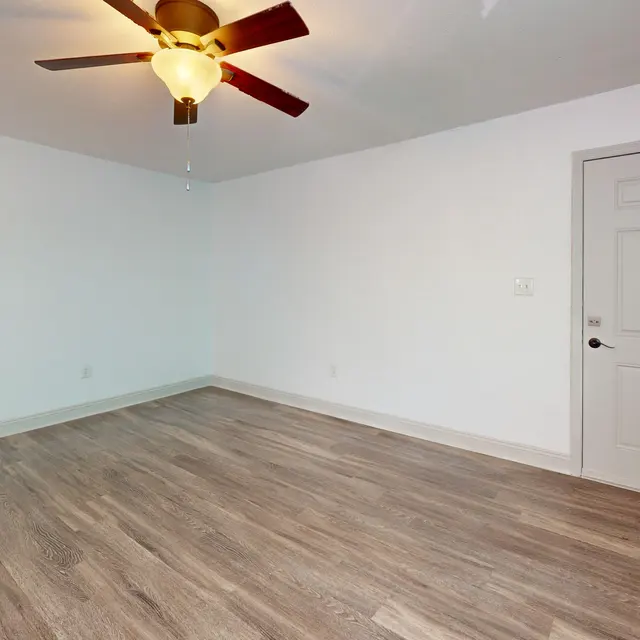 A spacious, empty room with light wooden flooring, a white ceiling, and a ceiling fan. There is a door on the right side leading to another area, and the walls are painted white.